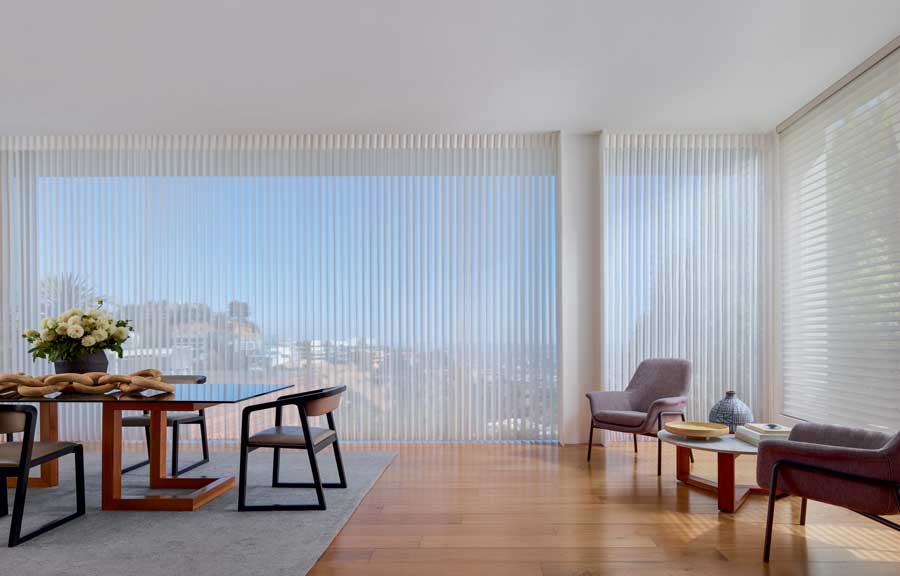 Large dining room with white window coverings. A dining table to the left and a conversation area with two chairs and a side table.