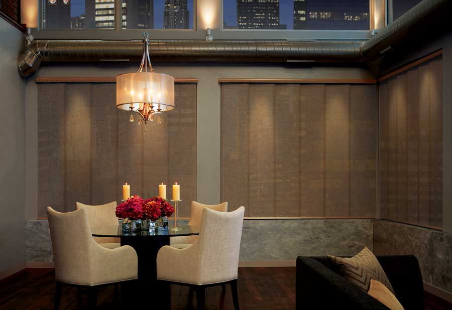 Tan window coverings covering the windows with a round dining table and chandelier and four fabric chairs surrounding the table.