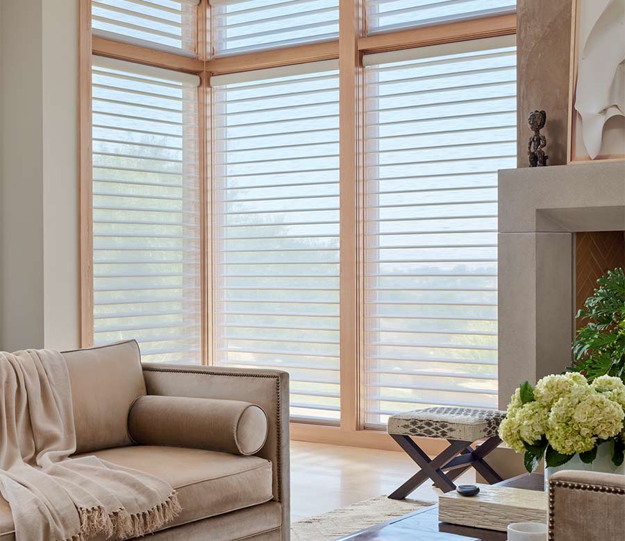 Silhouette® sheer window shades on floor-to-ceiling corner windows, creating a warm, diffused glow in a modern living room with sofa and fireplace